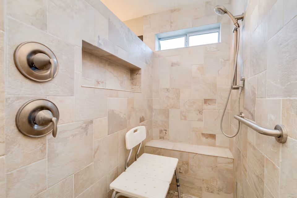 Tiled walk-in shower with a handheld showerhead, built-in bench, grab bar, and a white shower chair.