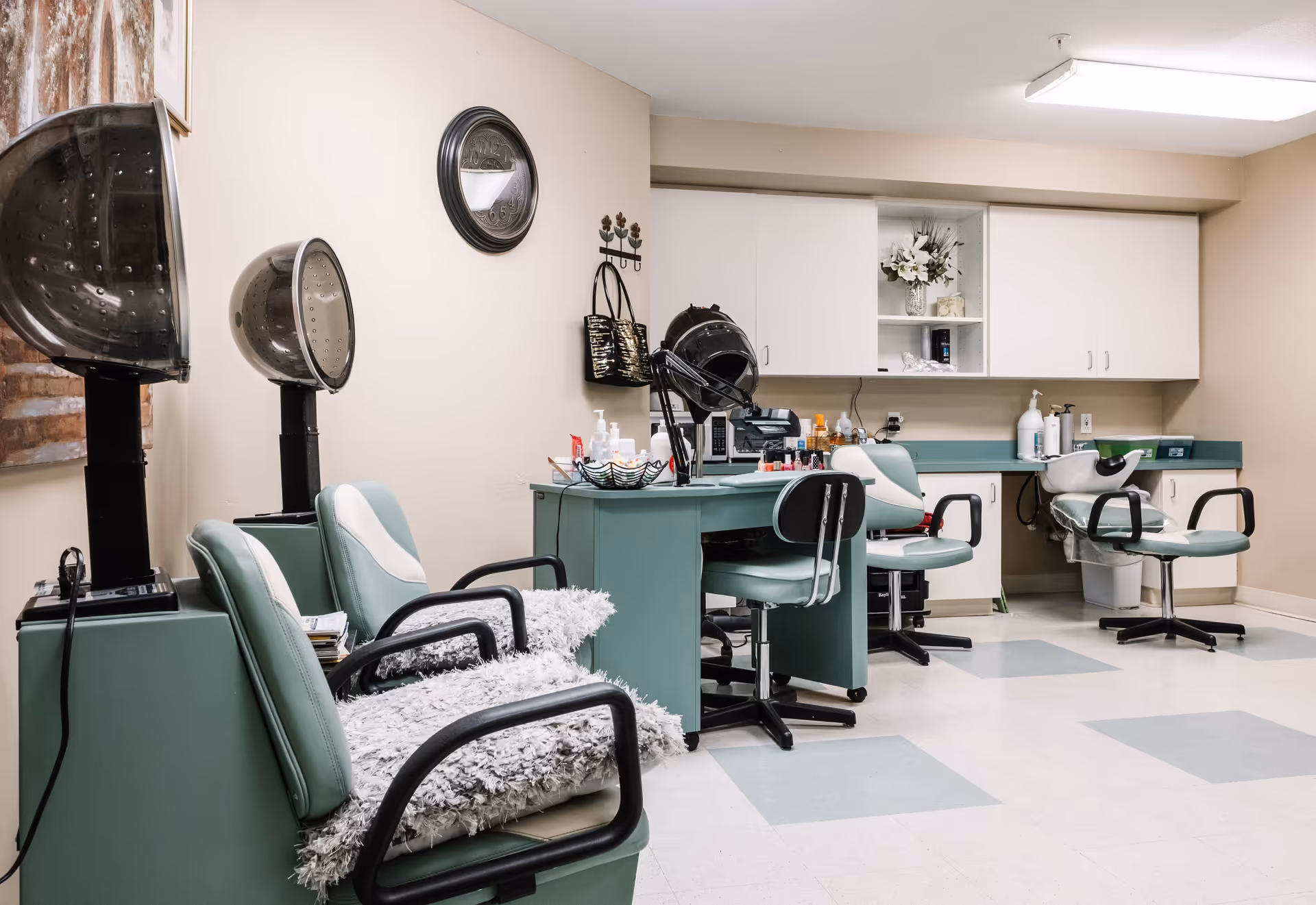 Interior view of a salon area in a senior living facility with green salon chairs, hair dryers, a workstation with beauty products, and white cabinets on the wall.