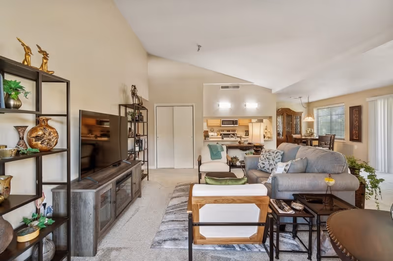 A bright and spacious living room with vaulted ceilings featuring a gray sofa, two armchairs, a TV on a wooden stand, decorative shelving with plants and vases, and a view into a kitchen and dining area with wooden furniture and large windows.