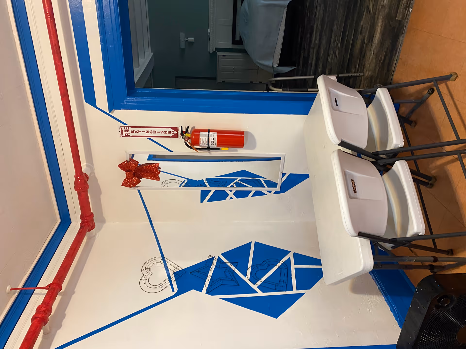 A small interior corner with a white folding table and two white folding chairs. The walls are decorated with blue geometric shapes and outlined heart and star designs. A rectangular mirror with a red glittery bow is mounted on the wall next to a fire extinguisher. The doorway with blue trim leads to a room with a bed and dark wood flooring.
