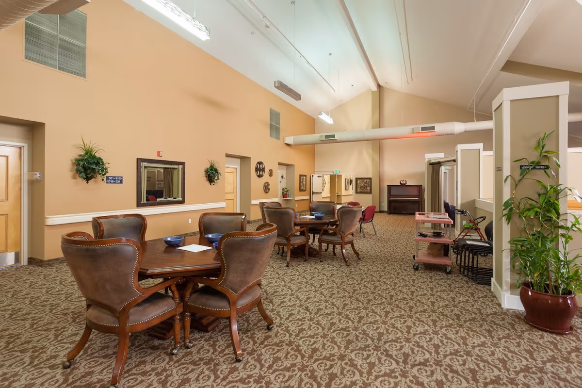 Spacious assisted-living common room with round wooden tables and chairs, patterned carpet, potted plants, and a piano in the background.