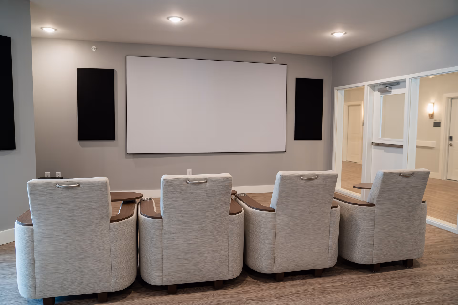 Four upholstered recliner chairs face a blank projection screen in a small media room.