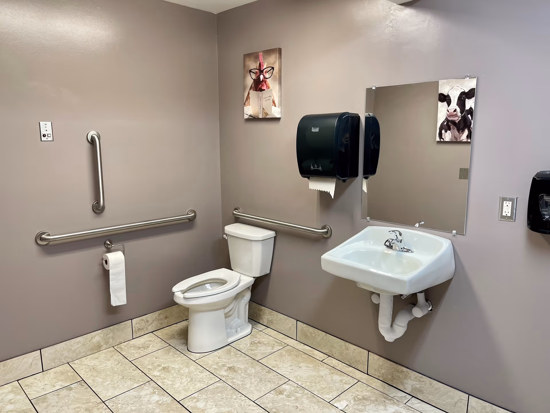 A clean and accessible bathroom with beige walls and tiled floor. The room features a white toilet with grab bars on the adjacent walls, a white wall-mounted sink with a faucet, a black paper towel dispenser, and a black soap dispenser. There are two small paintings on the walls, one of a chicken wearing glasses and reading a book, and another of a cow. A mirror is mounted above the sink.