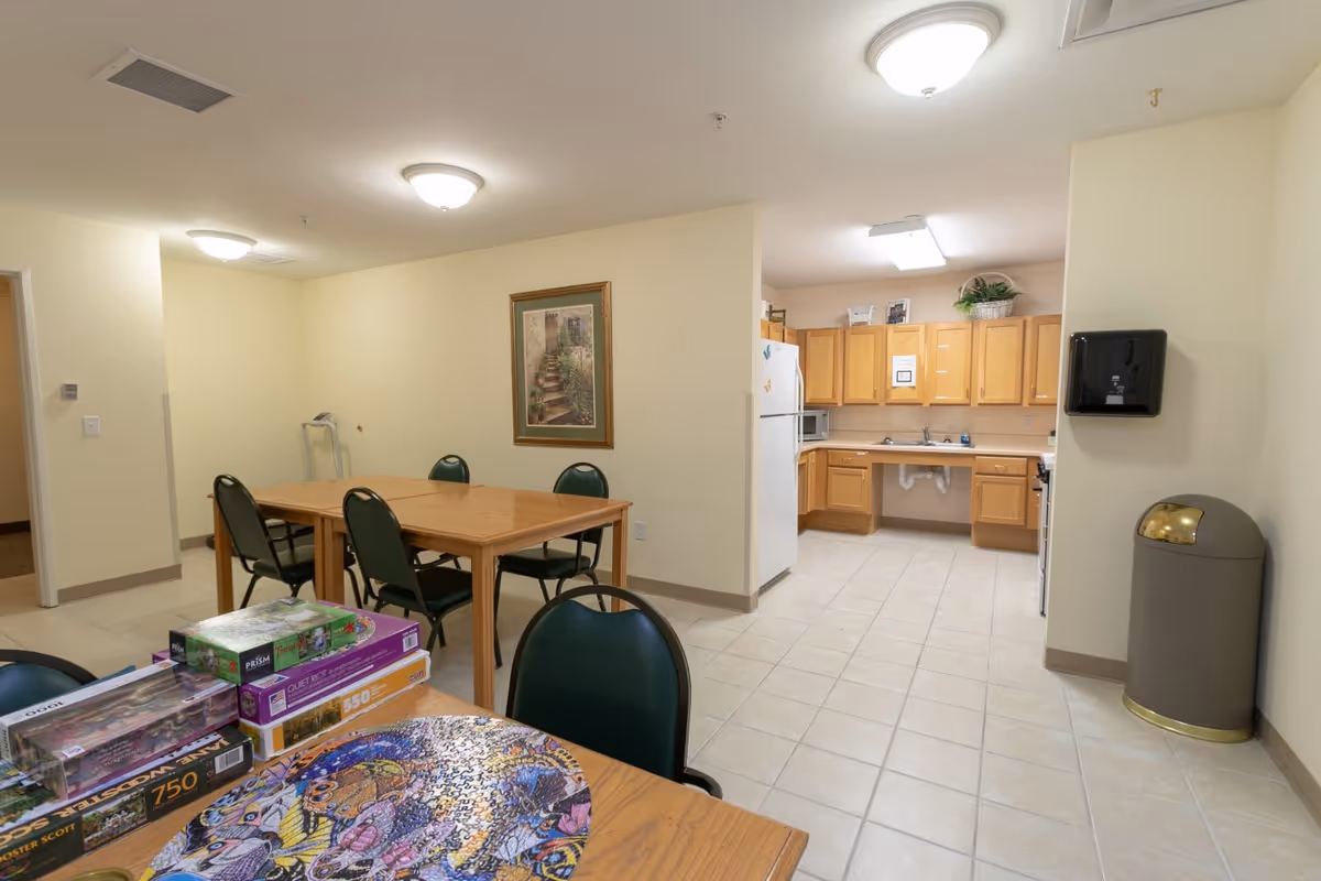 Interior view of a common area in a senior living facility showing a small kitchen with wooden cabinets, a white refrigerator, and a sink. In the foreground, there are tables with green chairs and several jigsaw puzzles on one of the tables. The walls are light-colored, and there is a framed picture hanging on the wall. The floor is tiled, and ceiling lights illuminate the room.