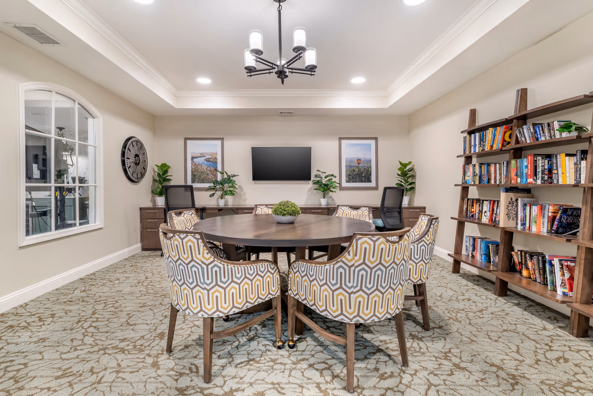 A well-lit meeting or activity room with a round wooden table surrounded by six patterned upholstered chairs. The room features a carpeted floor, a wall-mounted flat-screen TV, two framed landscape pictures, two potted plants on a wooden cabinet, a large wall clock, and a wooden bookshelf filled with books.