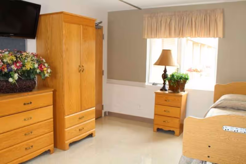 A simple, clean bedroom with light wood furniture including a dresser, wardrobe, and nightstand. The nightstand holds a lamp and a potted plant. There is a bed partially visible on the right side of the image. A window with beige curtains allows natural light into the room.