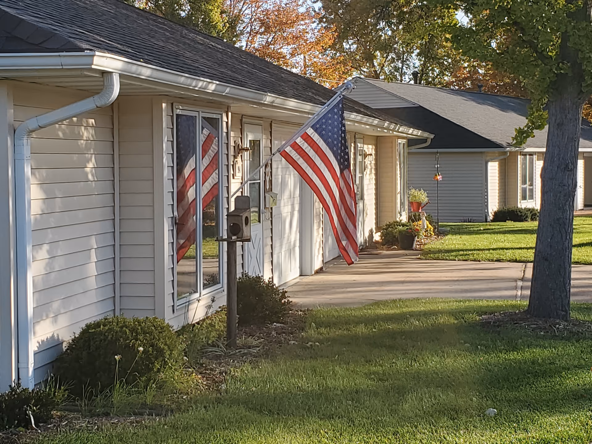 Exterior view of a single-story residential building with beige siding, a black shingled roof, and an American flag mounted near the front door. There is a small garden with bushes and a birdhouse in front of the building, and a tree on the right side. The scene is set in a well-maintained grassy area with other similar buildings visible in the background.