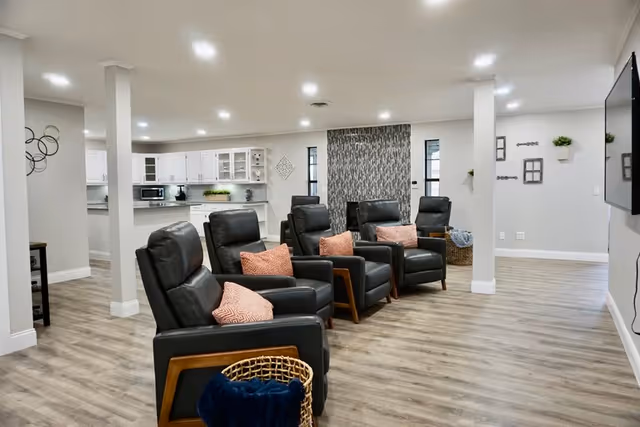 Bright communal lounge with rows of leather recliners and pink throw pillows facing a wall-mounted TV and an open kitchenette in the background.