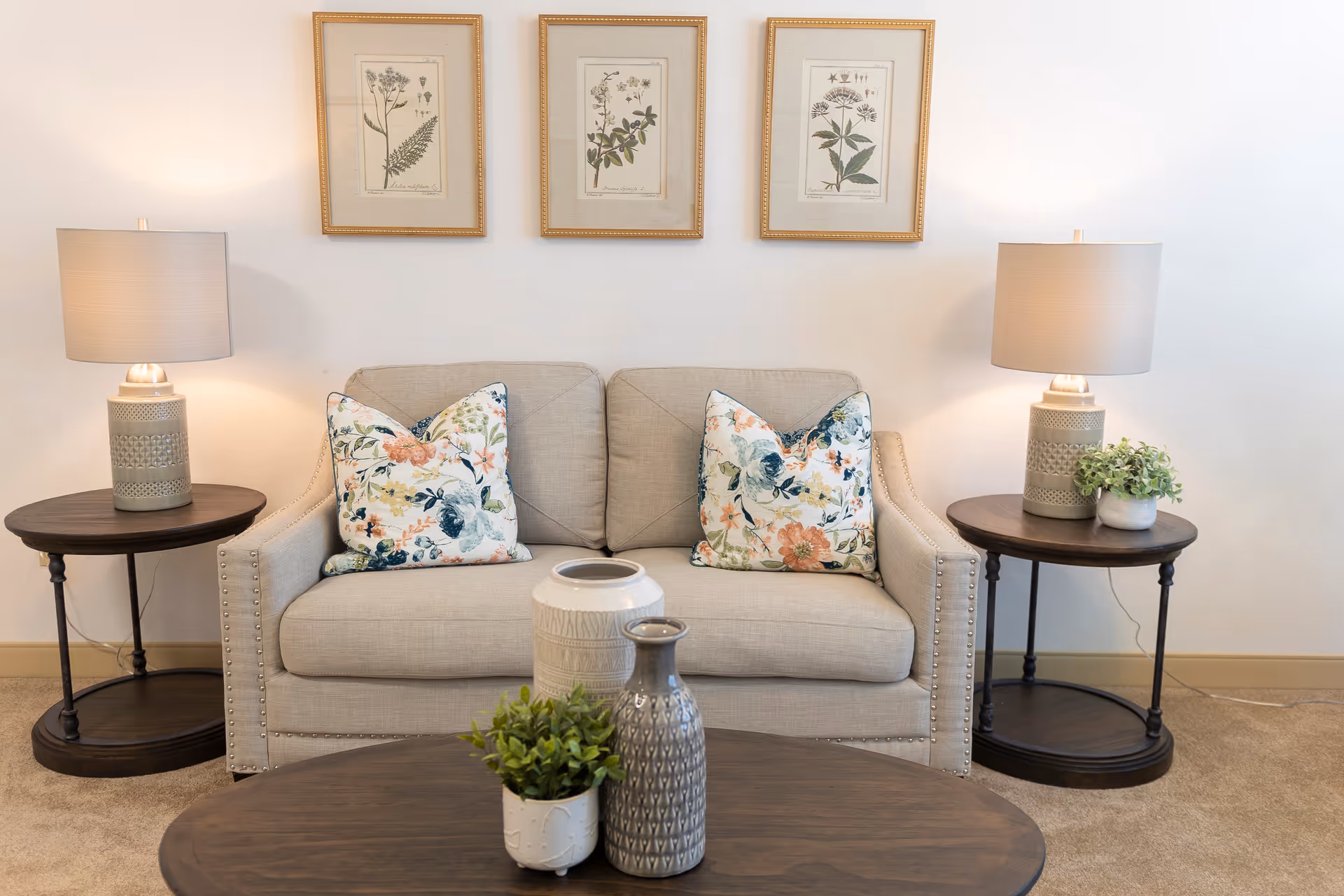 A cozy living room area with a beige loveseat adorned with two floral-patterned pillows. On either side of the loveseat are dark wooden side tables, each holding a beige lamp with a white lampshade. Above the loveseat, three framed botanical prints are hung on the wall. In front of the loveseat is a dark wooden coffee table with three decorative vases and a small potted plant. The room has a neutral color palette with soft lighting.