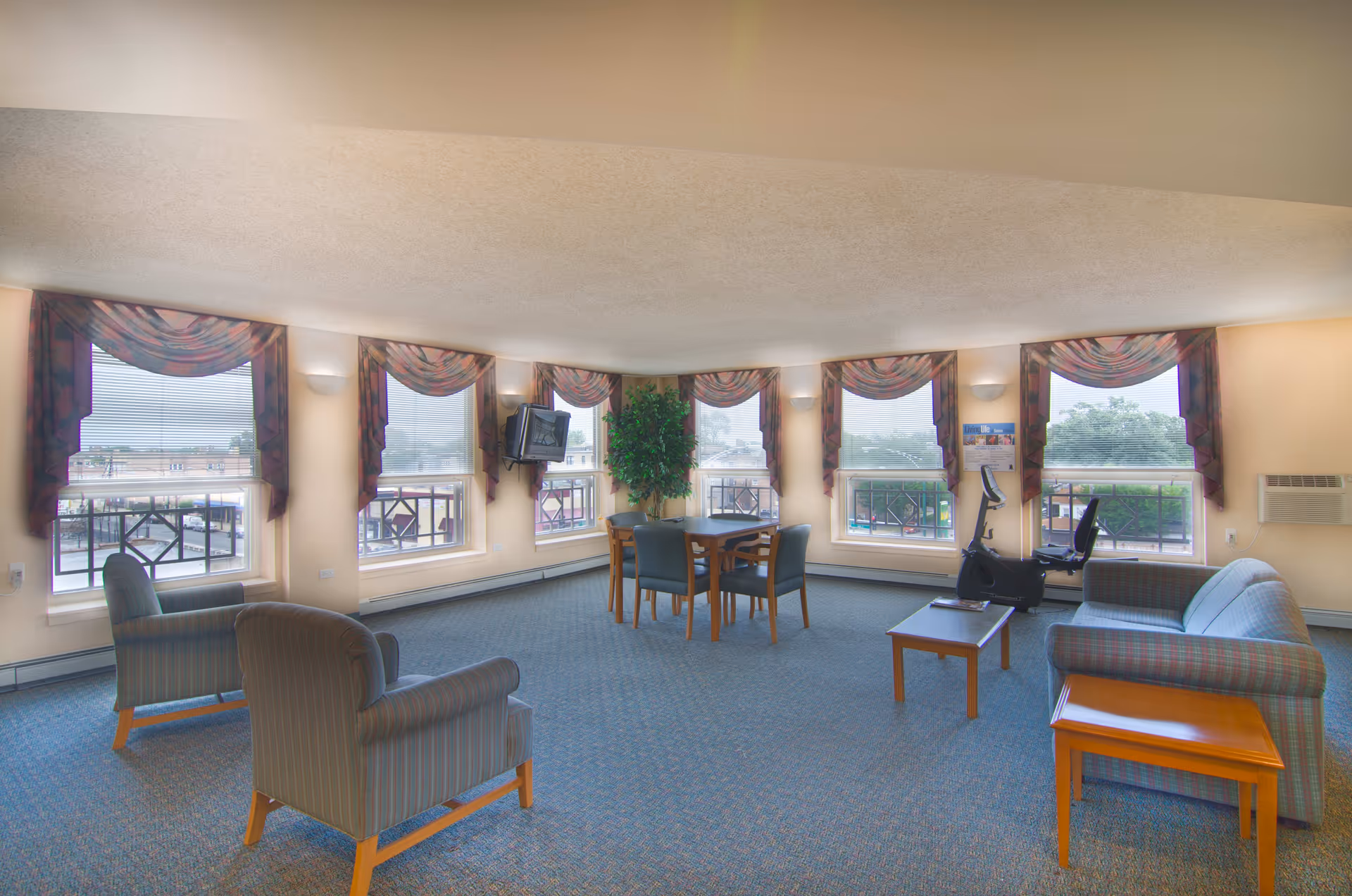 A spacious senior living facility common room with multiple large windows covered by patterned valances. The room features two armchairs, a sofa, a coffee table, a side table, a dining table with four chairs, a stationary exercise bike, a wall-mounted TV, and a potted plant in the corner. The carpet is blue with a subtle pattern, and the walls are painted a light beige color.