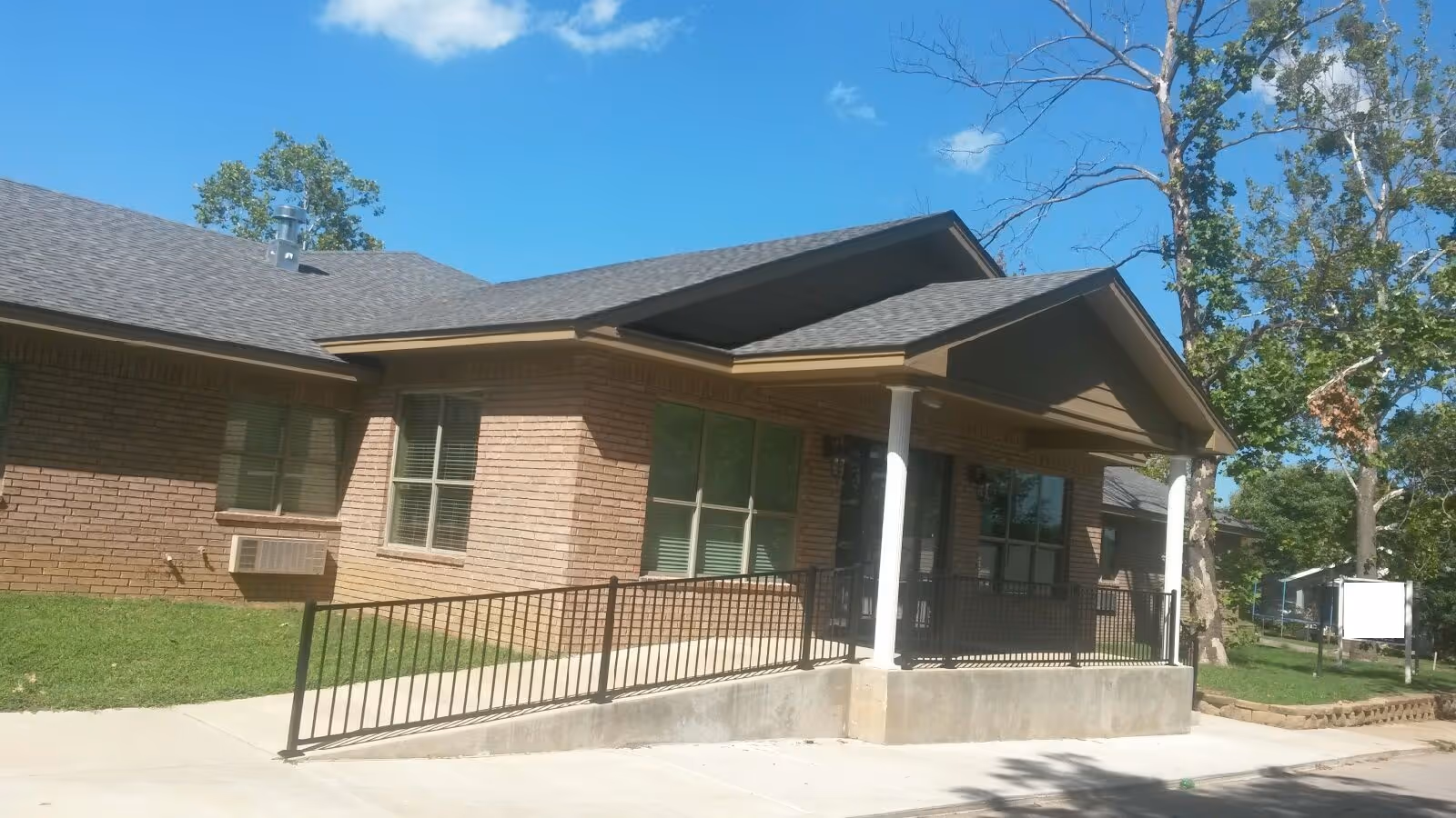 Exterior view of a single-story brick building with a covered entrance supported by white columns and a black metal railing along a concrete ramp. The building is surrounded by green grass and trees under a clear blue sky.