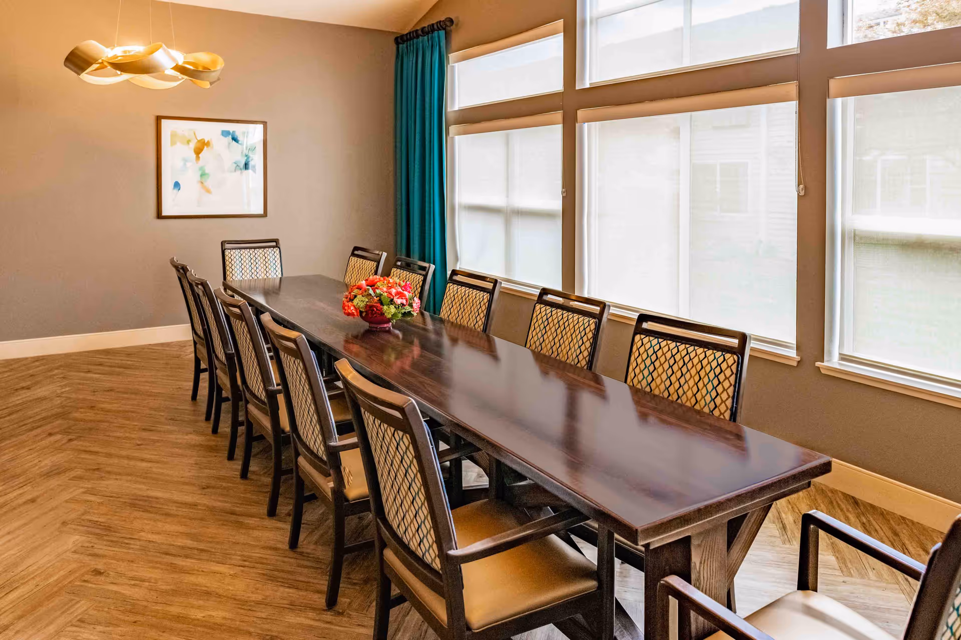 A long wooden dining table with twelve chairs arranged around it in a well-lit room. The table has a floral centerpiece. Large windows with white blinds and teal curtains allow natural light to fill the room. A modern light fixture hangs from the ceiling, and a framed abstract painting is on the wall.