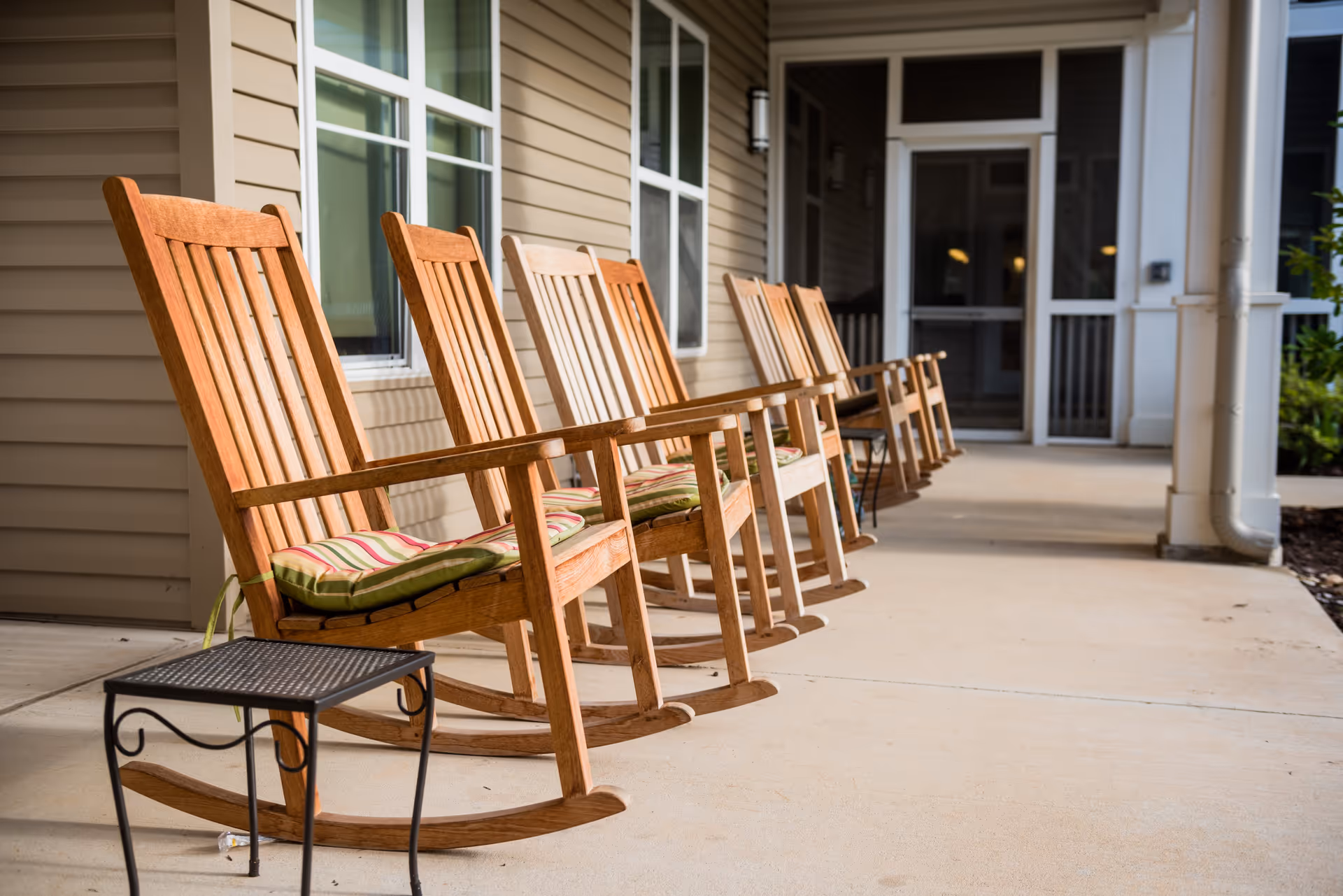 A row of wooden rocking chairs with striped cushions lined up on a covered porch outside a building with beige siding and white-framed windows and doors.