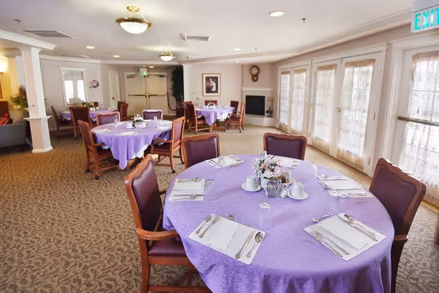 A dining room in an assisted living facility with round tables covered in purple tablecloths. Each table is set with placemats, silverware, napkins, glasses, and small floral centerpieces. The room has carpeted floors, large windows with sheer curtains, and a fireplace in the background.