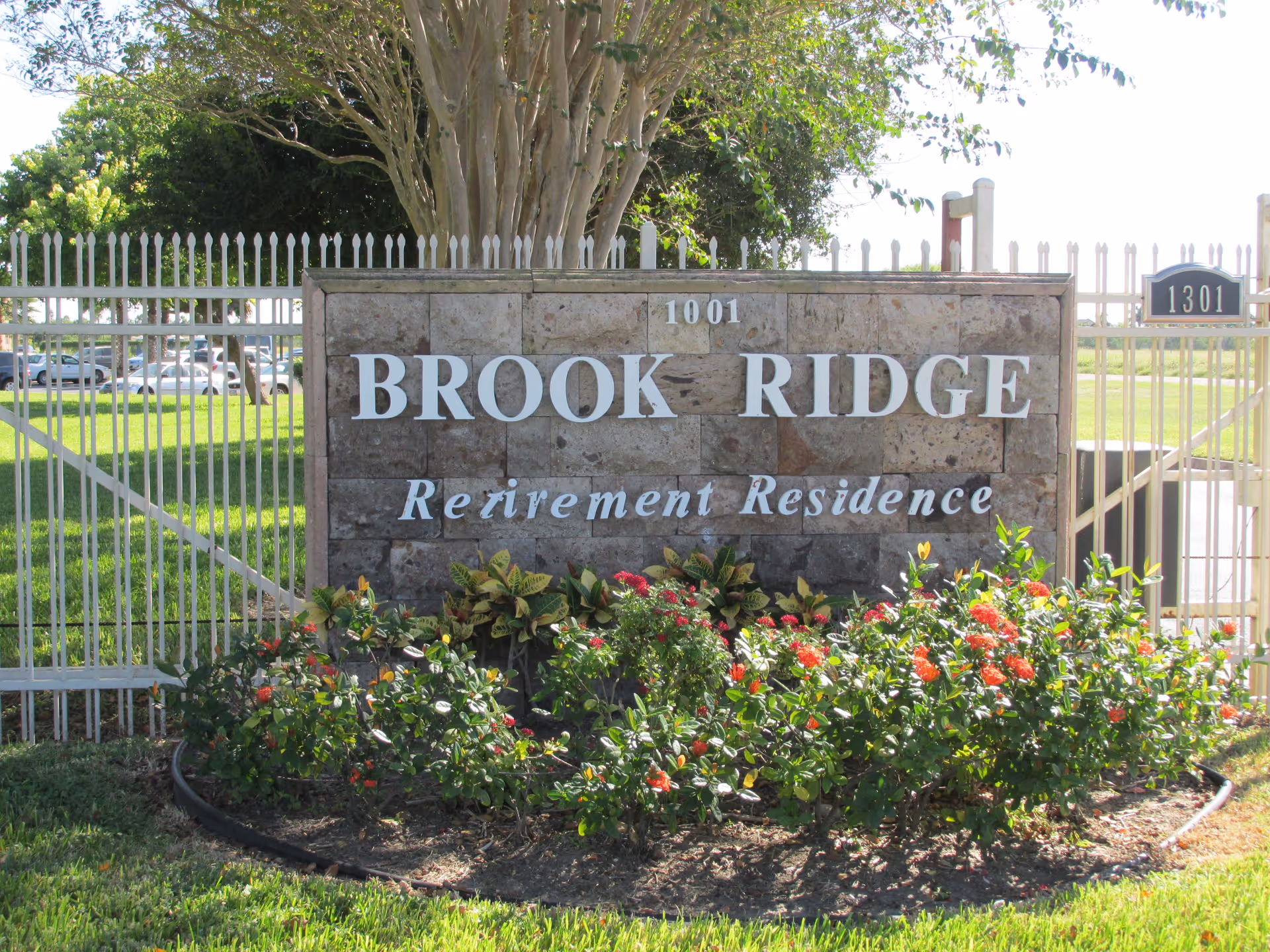 Stone sign at the entrance of Brook Ridge Retirement Residence with white metal fence and green lawn in the background, surrounded by flowering plants.