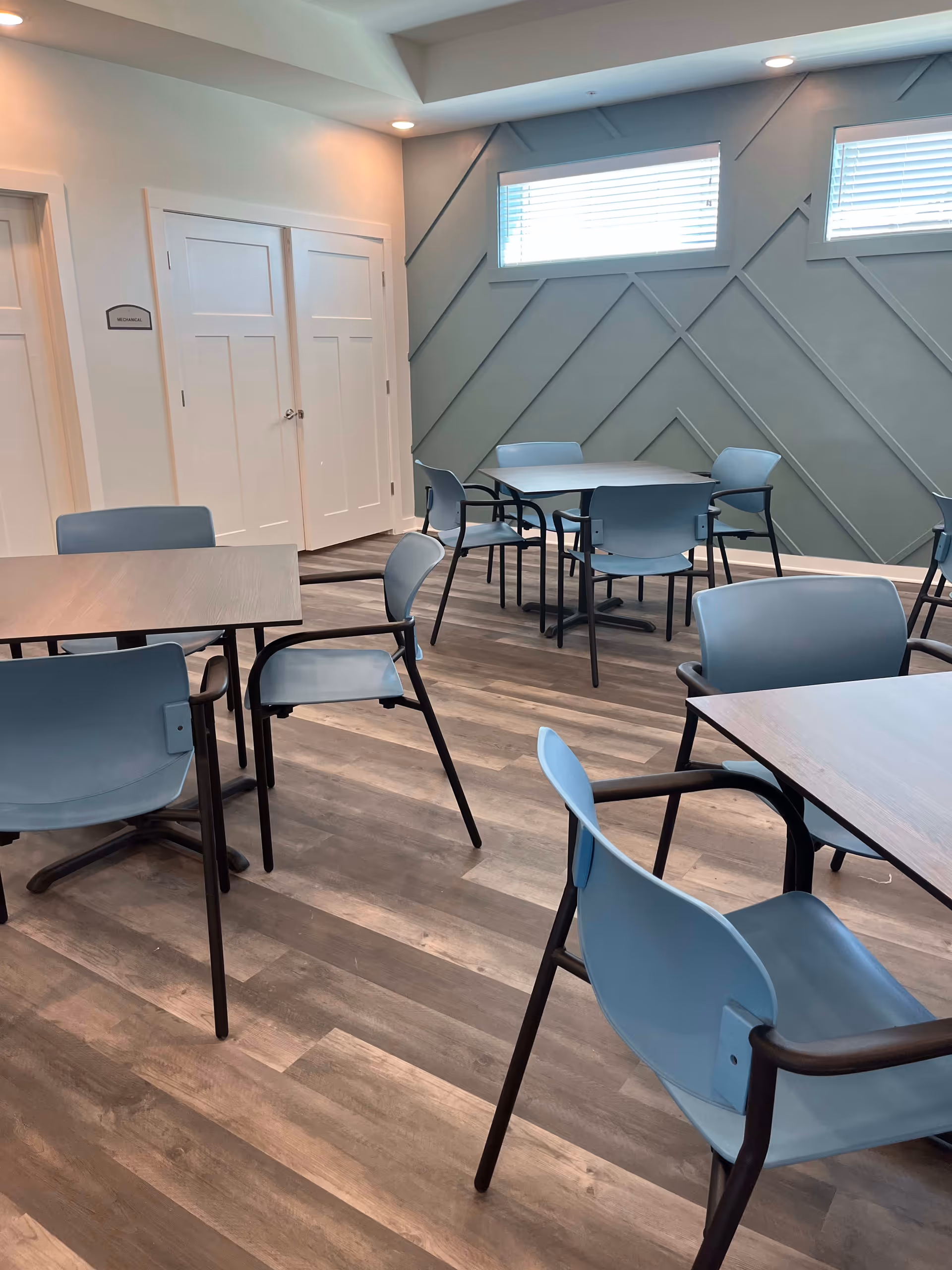 A bright dining area with several tables and blue chairs arranged on a wood-patterned floor. The walls are light-colored with a decorative geometric design on one side and two small windows letting in natural light. There is a door labeled 'Mechanical' on the left side of the room.