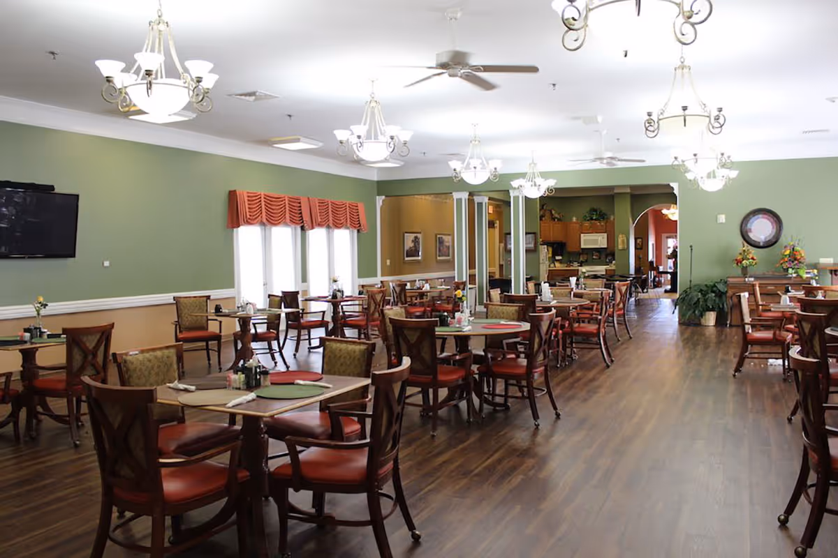 A spacious senior living dining room with multiple wooden tables and chairs arranged neatly. The walls are painted green with a beige lower section, and there are several chandeliers and ceiling fans hanging from the white ceiling. A flat-screen TV is mounted on one wall, and windows with red valances allow natural light to enter. The room has a warm, inviting atmosphere with wooden flooring and decorative plants and flowers.