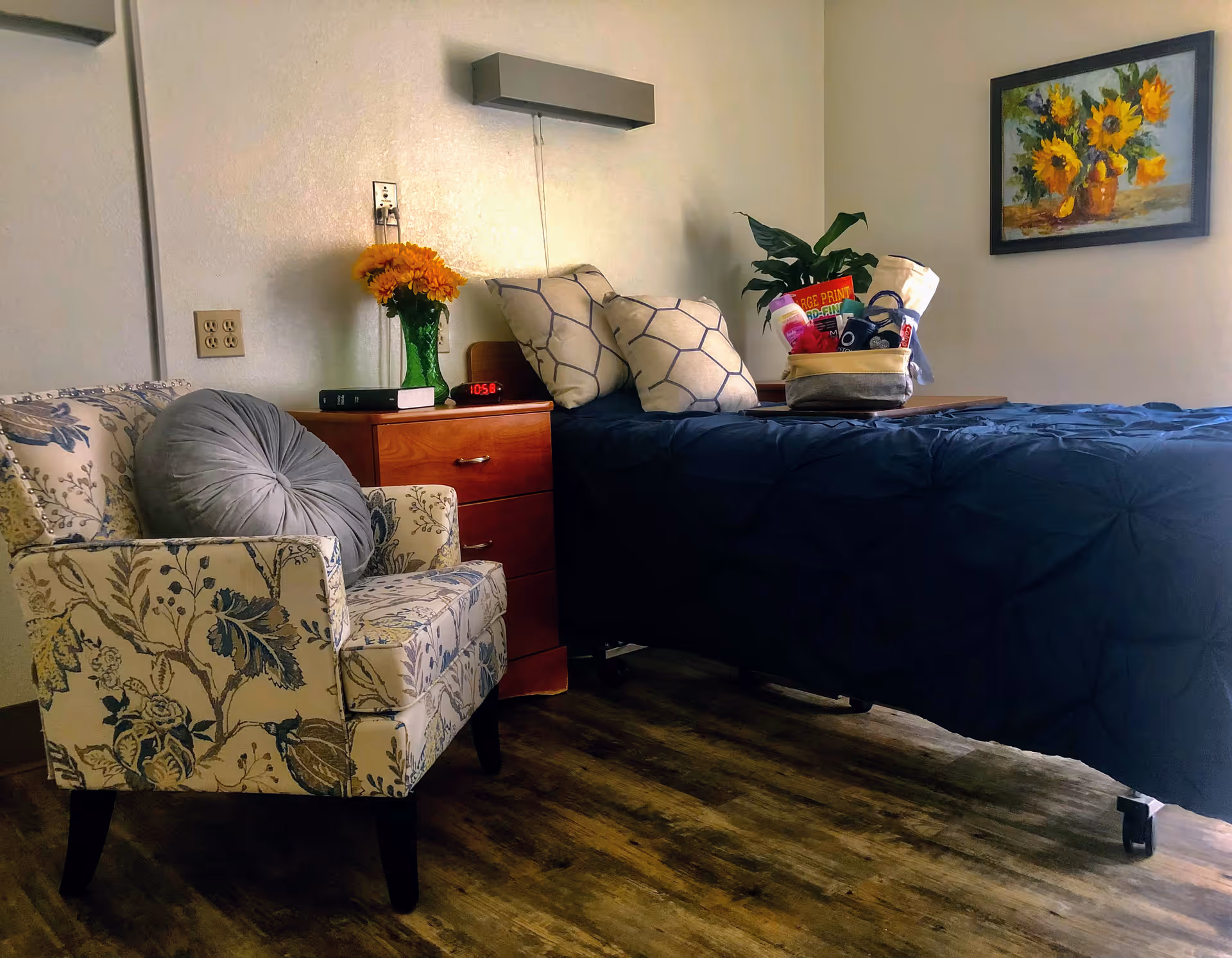 A cozy senior living bedroom with a floral armchair, wooden nightstand topped with flowers and an alarm clock, and a bed with a dark blue comforter and pillows.