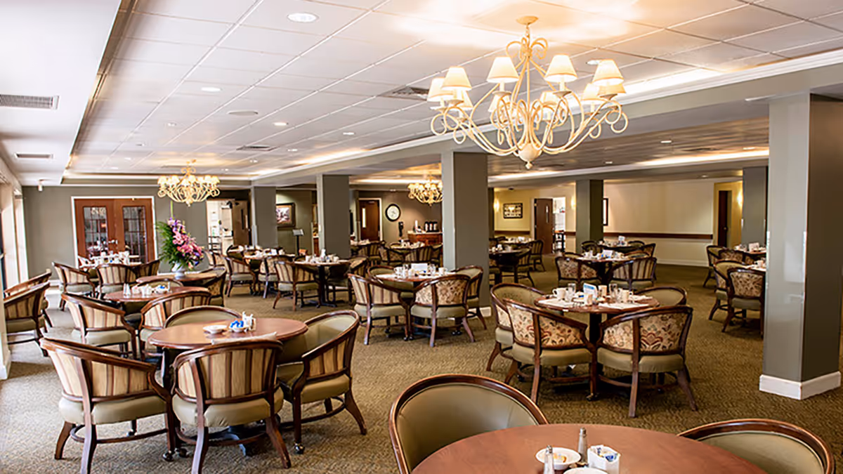 A spacious dining room with multiple round wooden tables surrounded by cushioned chairs. The room is well-lit with elegant chandeliers hanging from the ceiling. The tables are set with cups, saucers, and small condiment containers. The walls are painted in neutral tones, and there are large windows on one side letting in natural light.