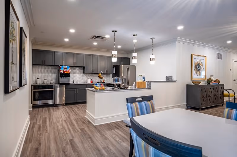 Modern kitchen and dining area in a senior living facility with gray cabinets, stainless steel appliances, a kitchen island with hanging pendant lights, and a dining table with striped chairs. The room has wood flooring, framed artwork on the walls, and a sideboard with decorative items.