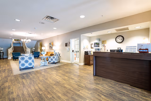 Interior view of a senior living facility lobby with a wooden reception desk on the right, a seating area with patterned armchairs and a small table on the left, and a staircase in the background. The floor is wooden, and the walls are light-colored with framed artwork and lighting fixtures.