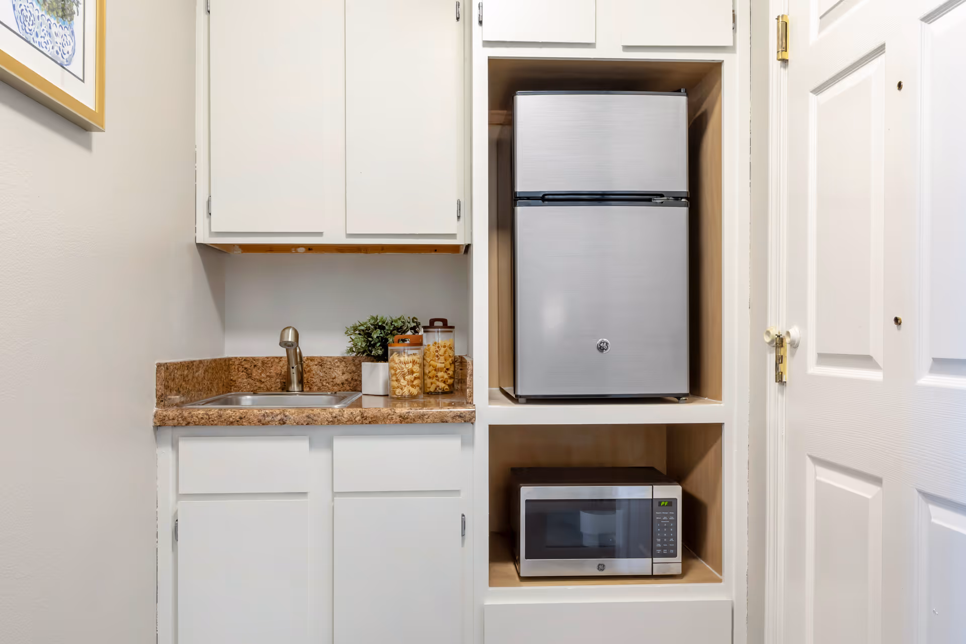 Small kitchenette area with a stainless steel mini refrigerator and microwave built into a white cabinet. There is a countertop with a sink, a faucet, two jars with snacks, and a small potted plant. A white door is visible on the right side and a framed picture hangs on the left wall.