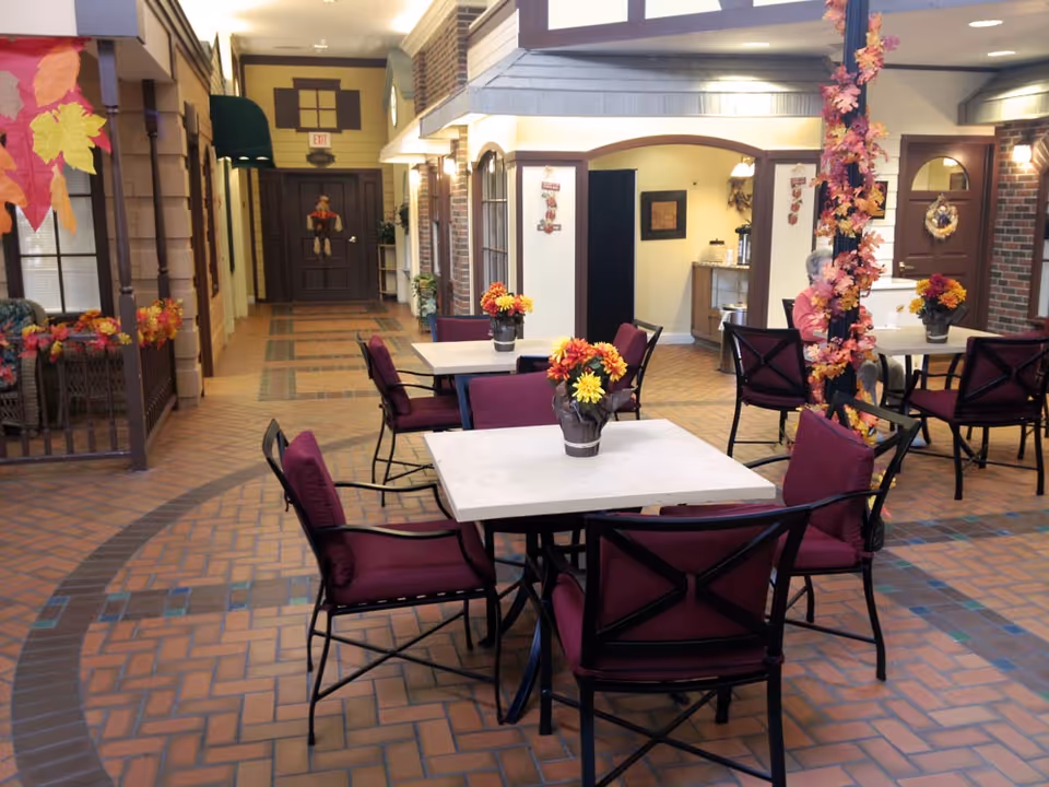 Indoor communal dining area with square tables, maroon chairs, and autumn decorations in a senior living facility.