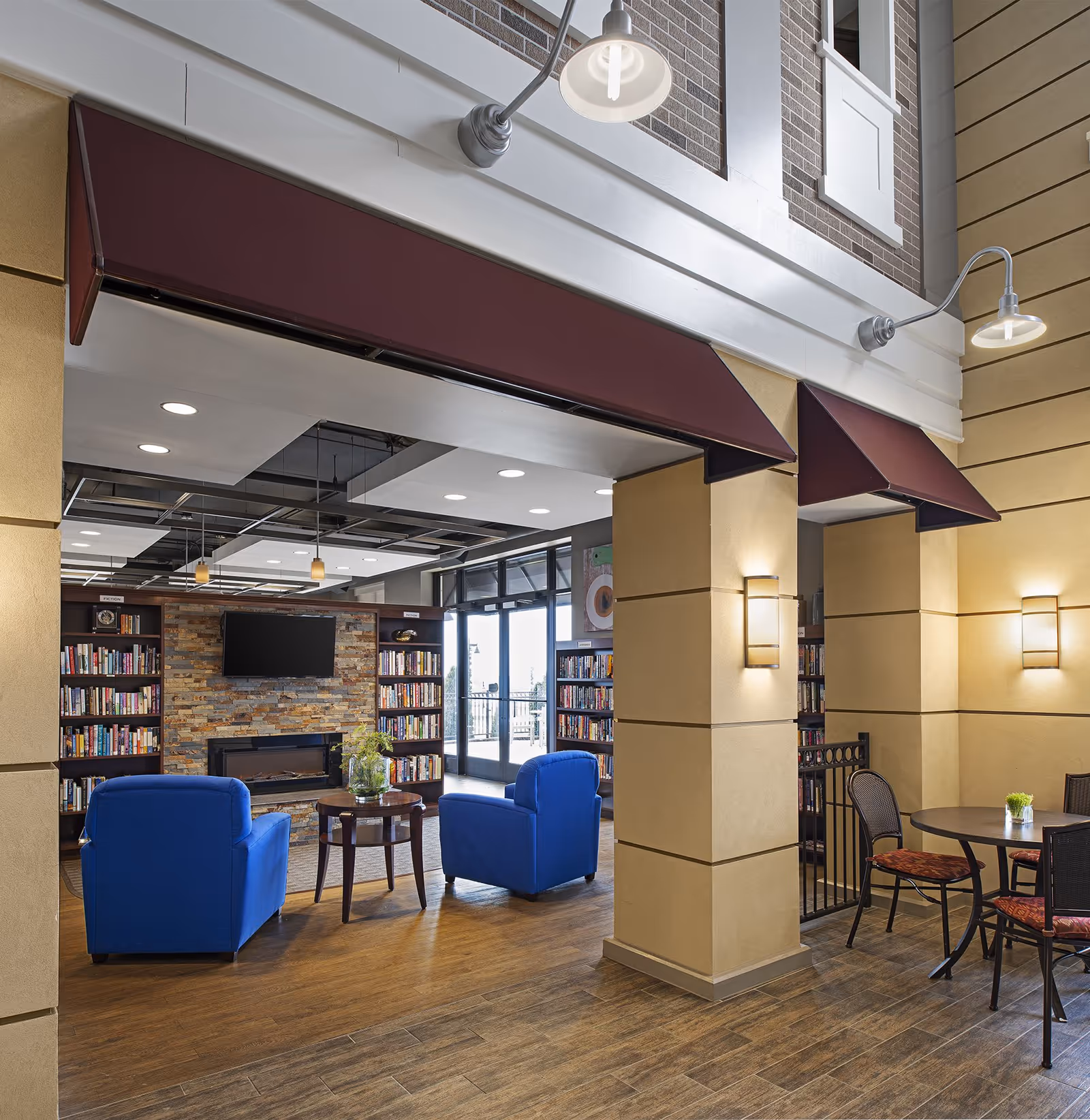 Interior view of a senior living facility lounge area featuring two blue armchairs facing a stone fireplace with a mounted flat-screen TV above it. Surrounding the fireplace are bookshelves filled with books. To the right, there is a small round table with four chairs, and the space is illuminated by wall-mounted lights and ceiling fixtures. The floor is wood and tile, and large windows provide natural light.