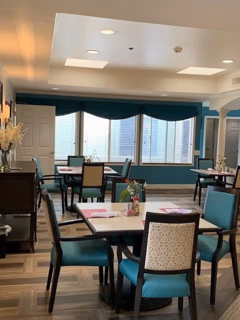 A dining room with several tables and chairs arranged neatly. Each table has place settings with napkins and small flower arrangements. Large windows with blinds allow natural light to fill the room. The walls are painted teal and white, and the floor has a patterned wood design.