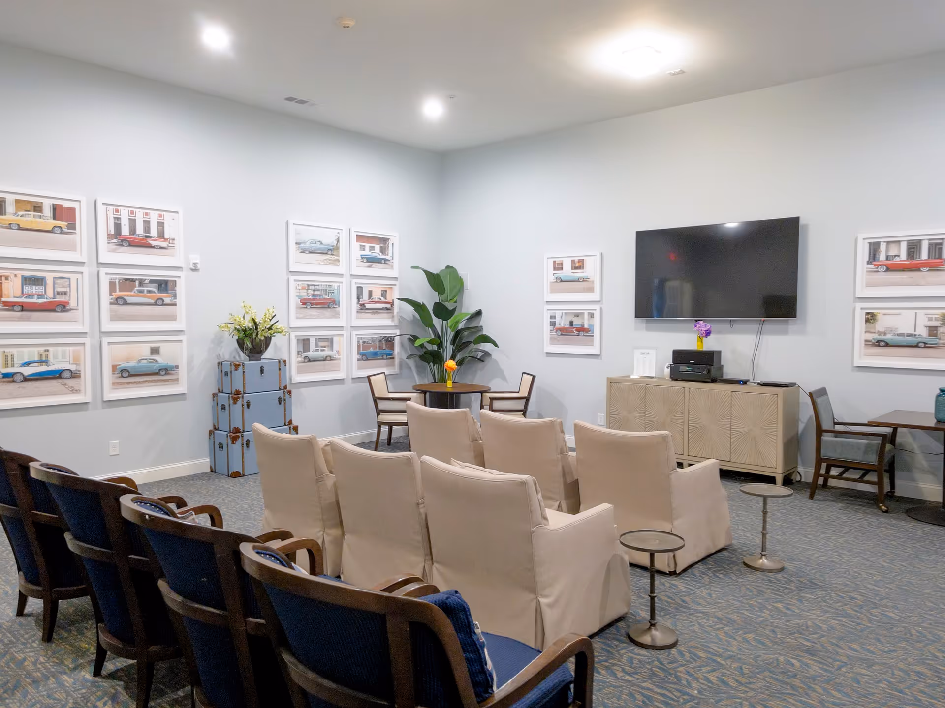 A cozy common area with rows of chairs facing a wall-mounted flat screen TV. The room features light blue walls adorned with framed pictures of vintage cars. There are small tables and chairs along the walls, a plant in the corner, and a cabinet beneath the TV with a small flower vase on top.