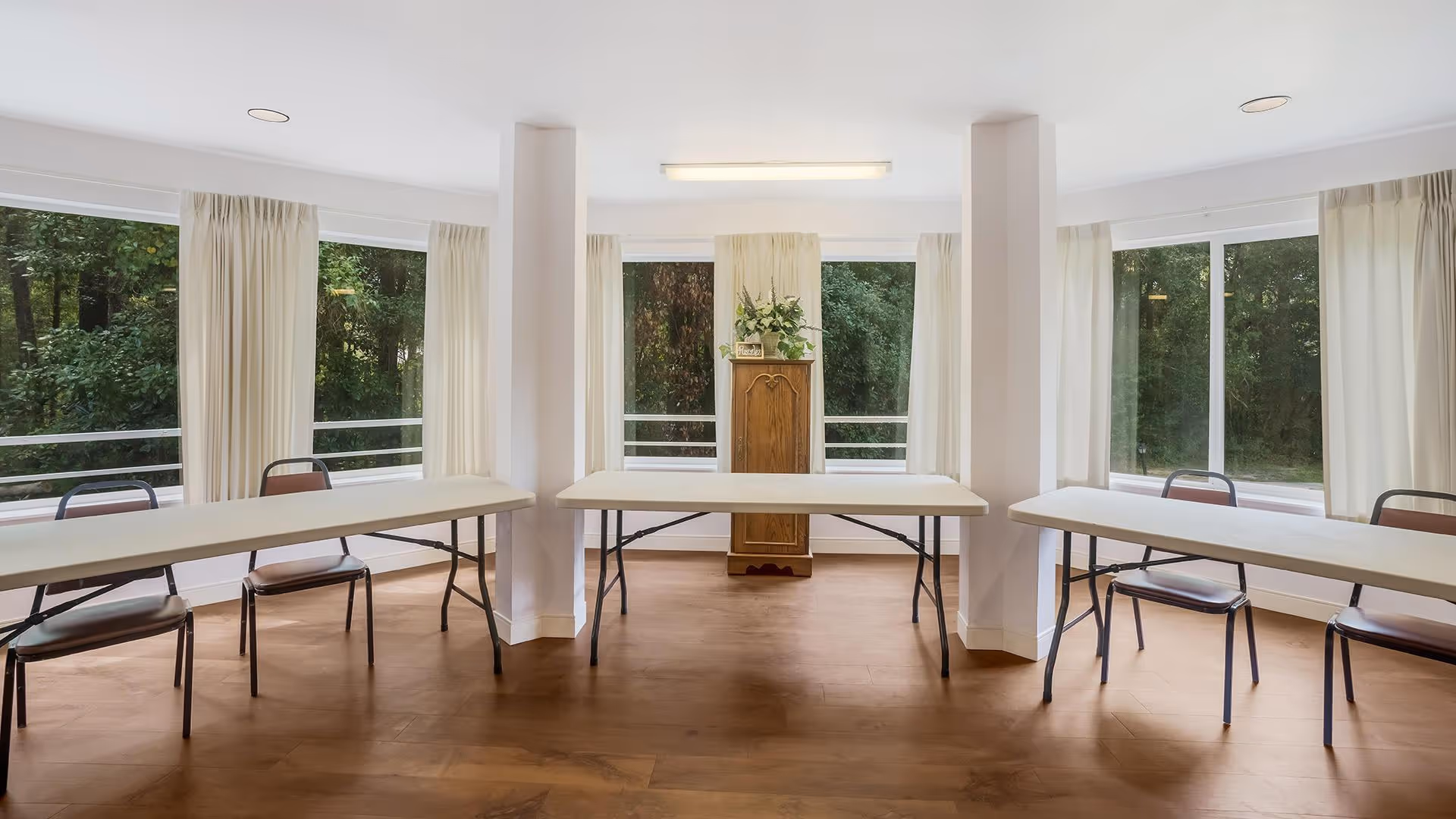 A bright room with large windows on three sides showing green trees outside. Inside, there are three long folding tables arranged in a U-shape with several chairs placed around them. In the center against the back wall, there is a wooden cabinet with a plant on top. The floor is wooden, and the walls are white with ceiling lights.