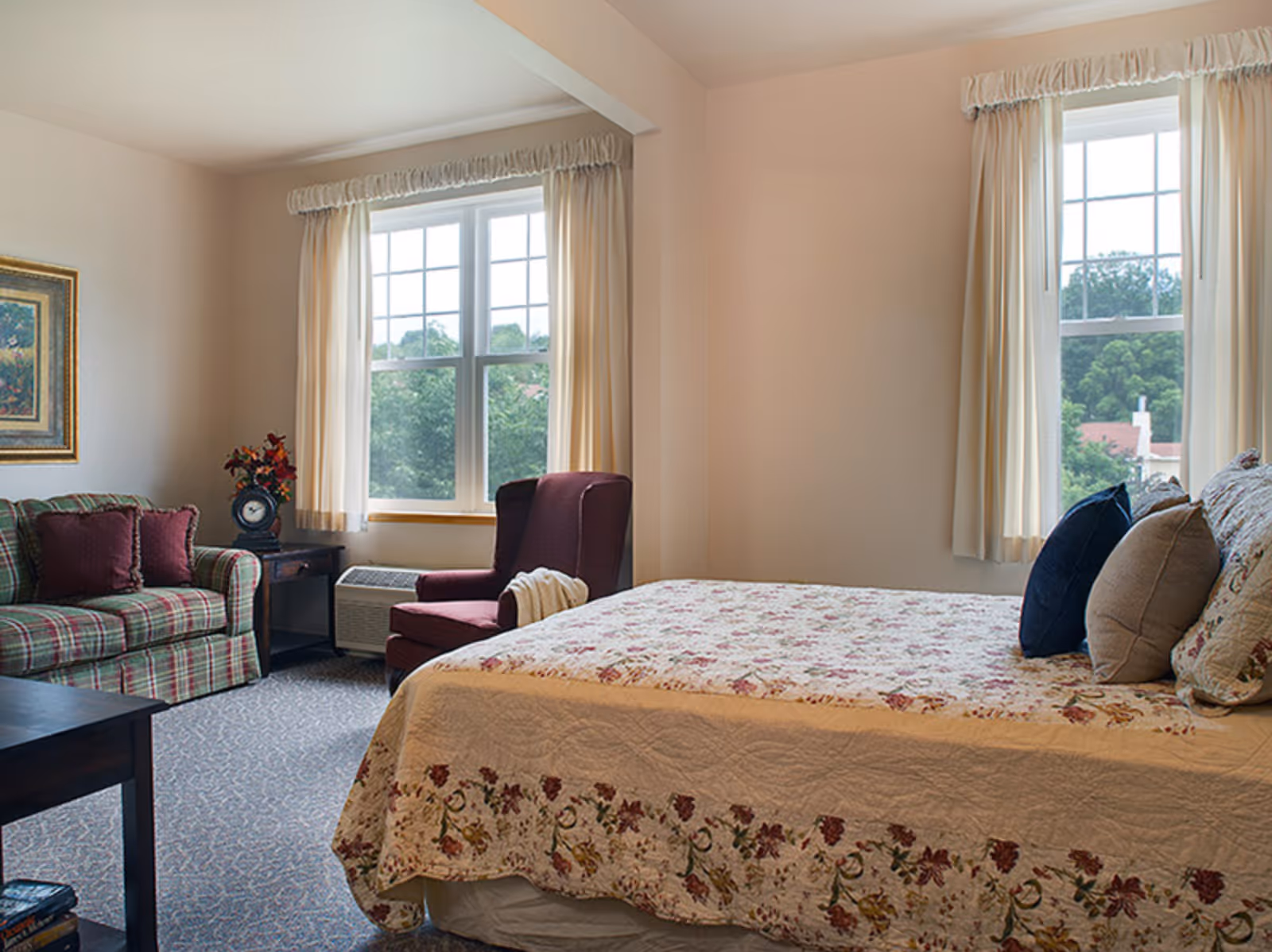 Bright furnished bedroom with a floral bedspread, a plaid sofa and armchair by two windows with cream curtains.