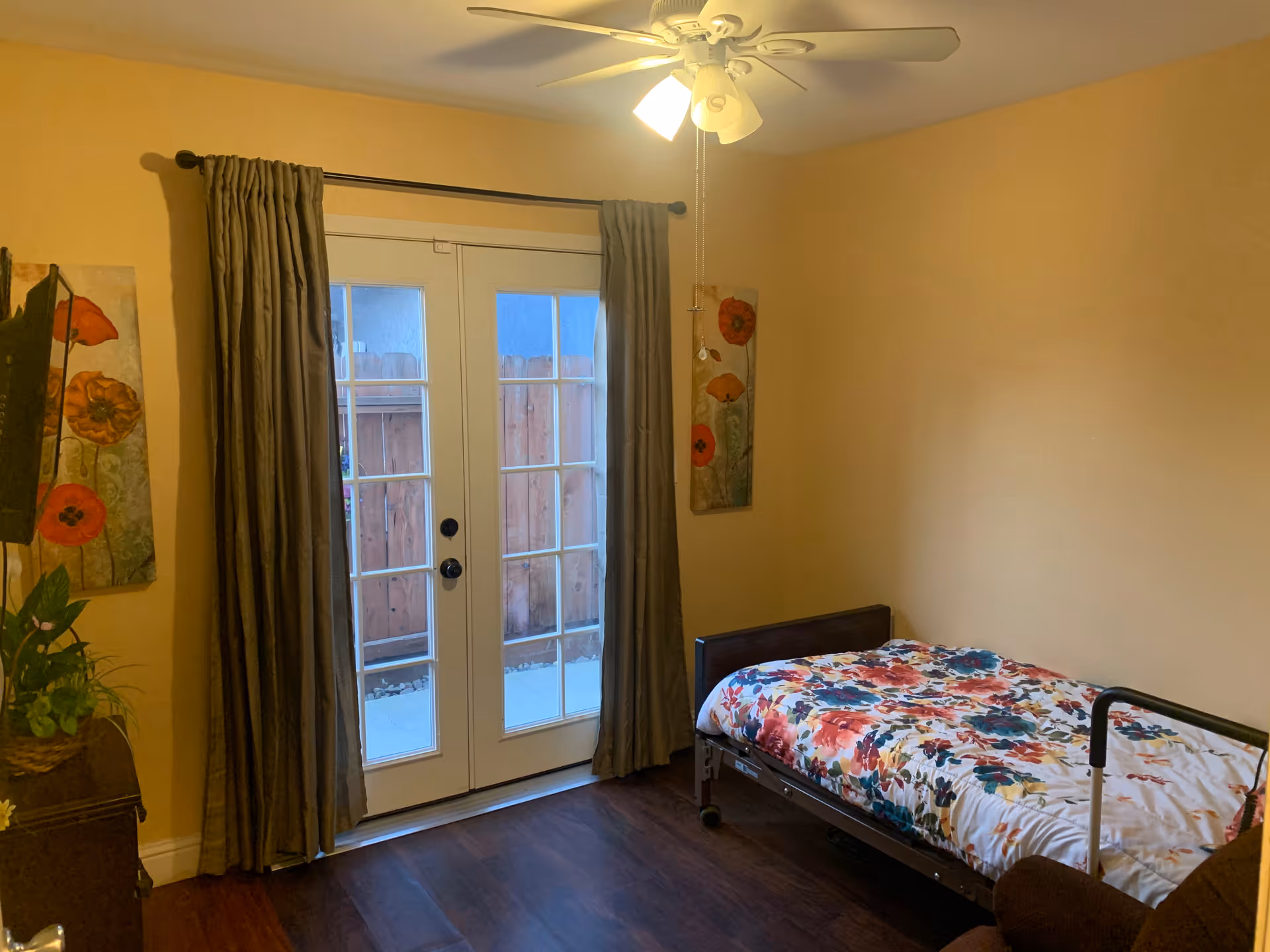 A small bedroom with a single bed covered in a colorful floral bedspread. The room has wooden flooring, beige walls, and a ceiling fan with lights. There are two paintings with red and orange flowers on the walls, and a set of French doors with brown curtains leading outside to a fenced area.