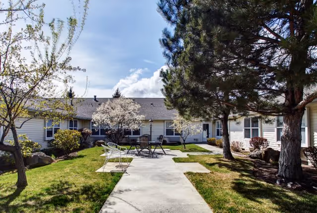 Outdoor courtyard area at Awbrey Place Assisted Living and Memory Care featuring a concrete walkway leading to a seating area with metal chairs and a table. The courtyard is surrounded by single-story building sections with windows, and there are trees and shrubs providing greenery and shade under a partly cloudy sky.
