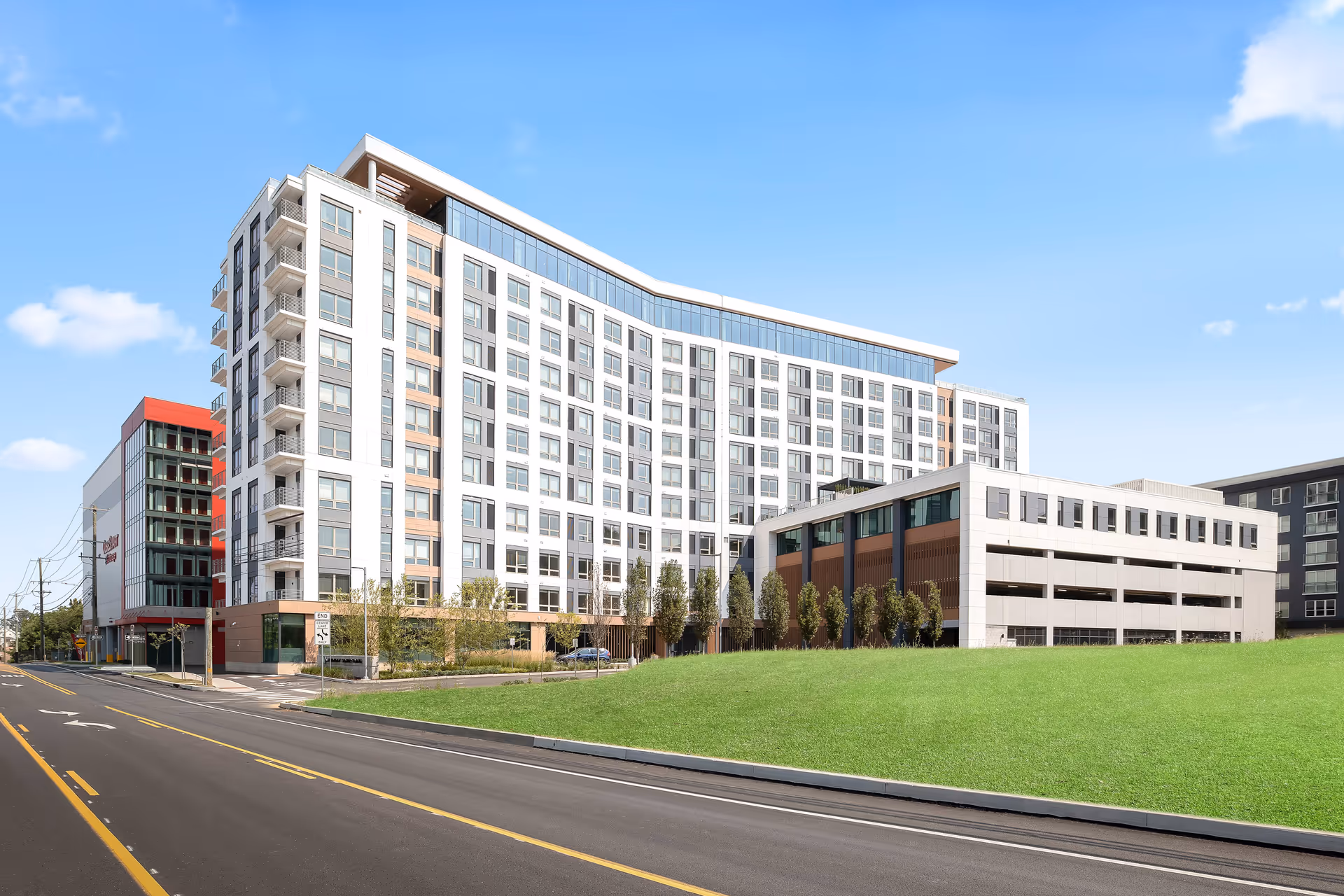 A large modern multi-story residential building with an attached parking garage, lawn, and road under a clear blue sky.