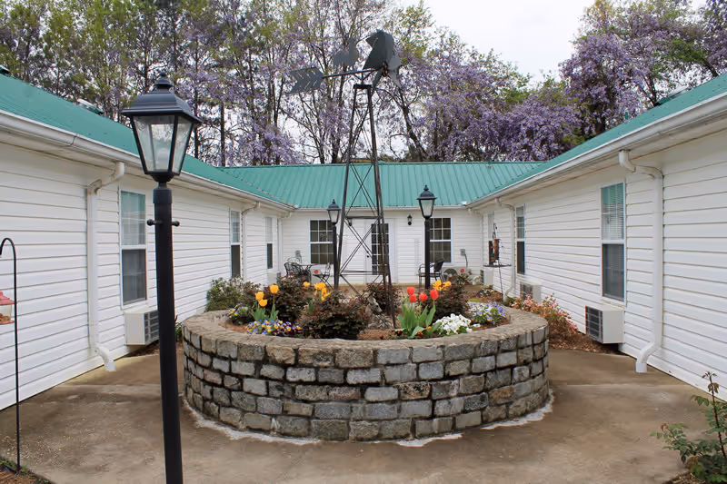 A courtyard area surrounded by white buildings with green roofs. In the center is a circular stone planter filled with colorful flowers and a small metal windmill structure. There are black lamp posts around the courtyard and trees with purple blossoms in the background.