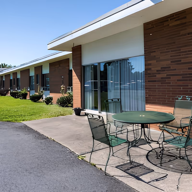 Outdoor patio with a round metal table and chairs in front of a single-story brick rehabilitation and nursing center with sliding glass doors and lawn.