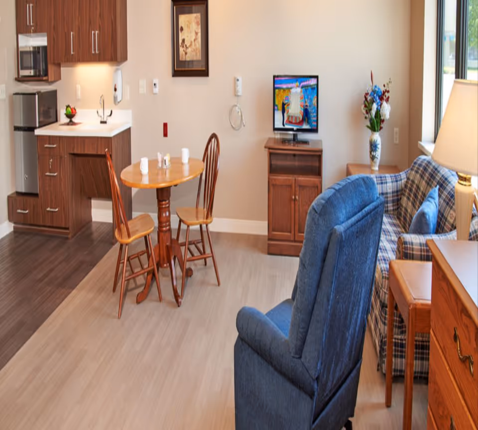 A cozy living area in a senior living facility featuring a small wooden dining table with two chairs, a kitchenette with wooden cabinets, a mini refrigerator, and a microwave. There is a blue upholstered recliner chair, a plaid sofa with blue cushions, a wooden TV stand with a flat-screen TV, a side table with a vase of flowers, and a lamp near a window letting in natural light.