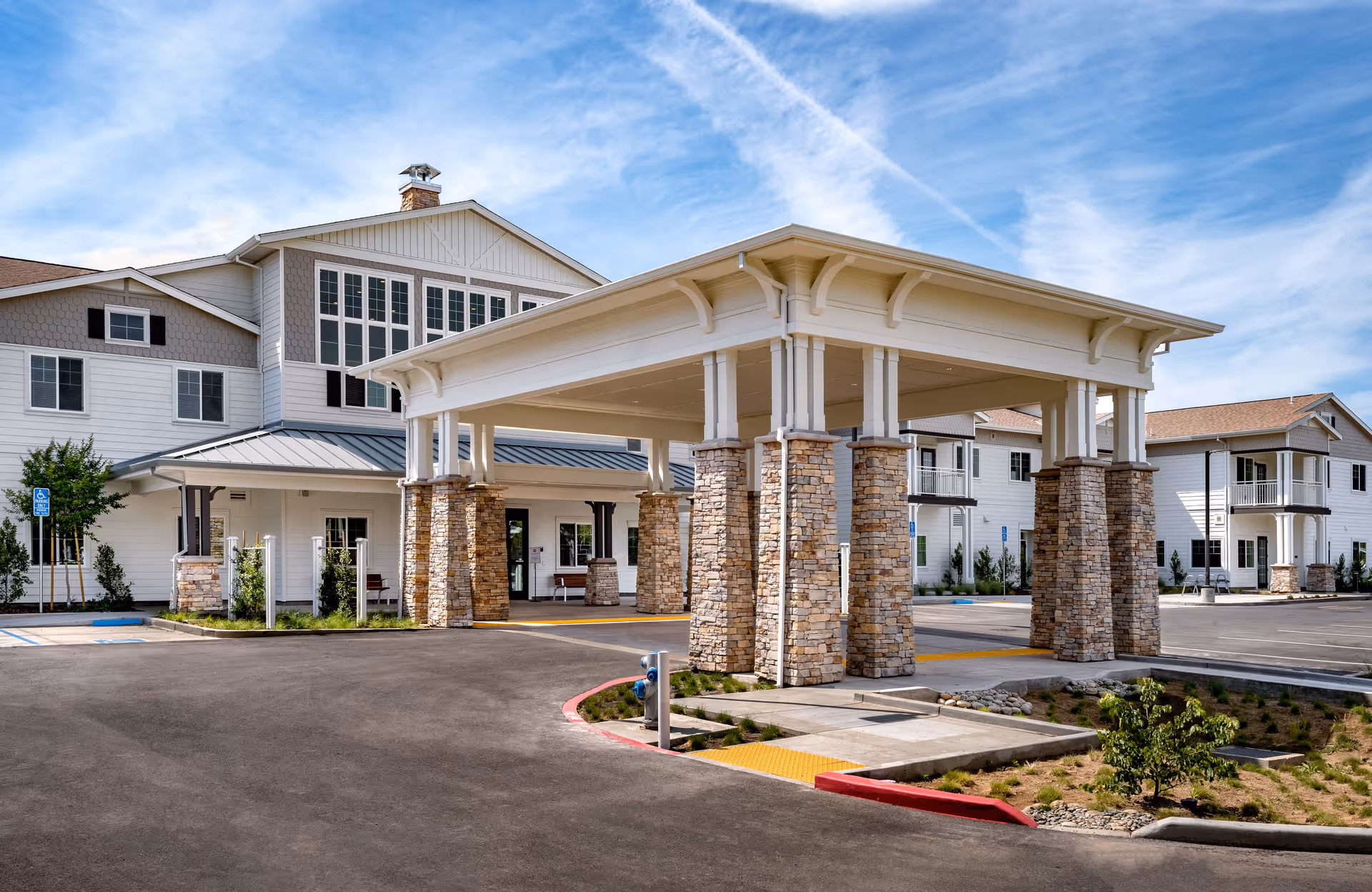 Exterior view of Clearwater at Sonoma Hills senior living facility showing a large covered entrance supported by stone pillars, with a multi-story white building in the background under a blue sky with some clouds.