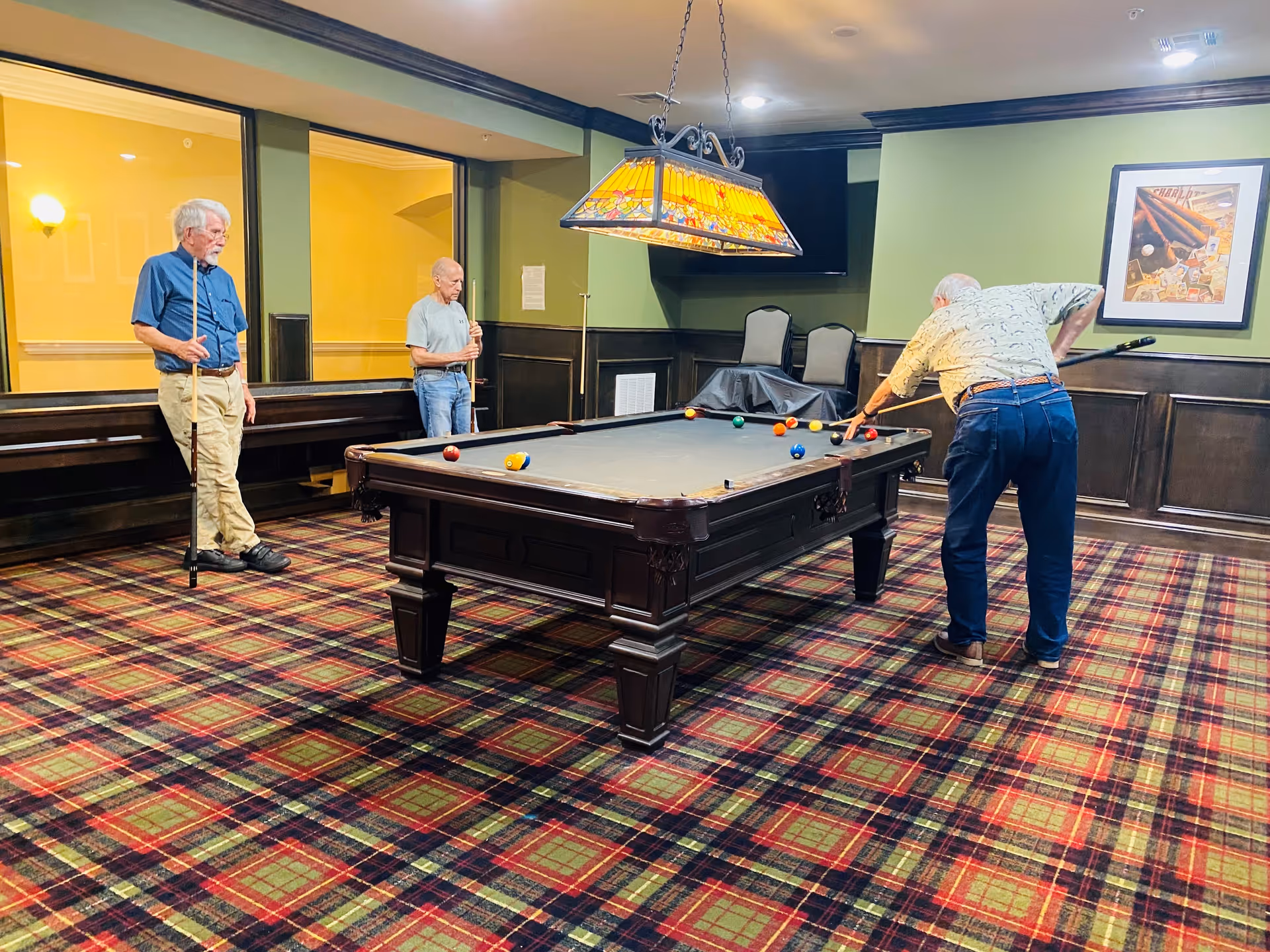 Three elderly men playing pool in a room with green walls, dark wood paneling, and a plaid carpet. One man is taking a shot while the other two watch, each holding a pool cue. A stained glass-style hanging light fixture is above the pool table, and a framed picture and a TV are mounted on the wall.