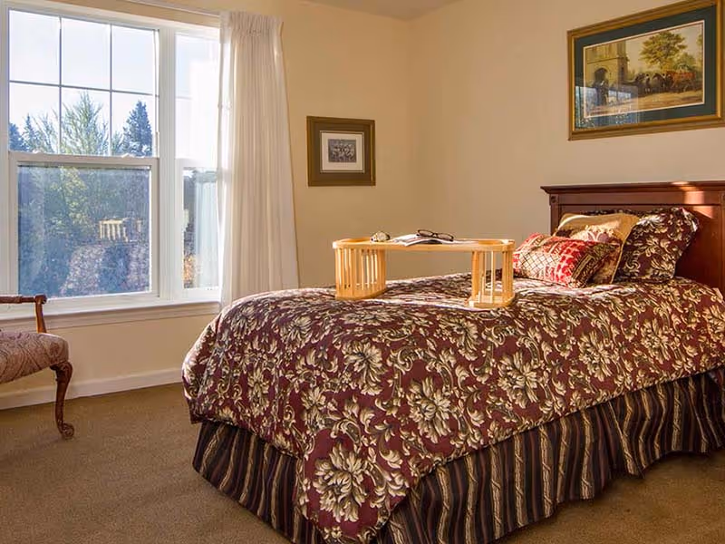 A cozy bedroom with a large window letting in natural light. The bed has a floral patterned bedspread in shades of burgundy and gold, with several decorative pillows. A wooden tray with a book and glasses rests on the bed. There is a framed picture on the wall above the bed and another smaller framed picture on the adjacent wall. A wooden chair with an upholstered seat is positioned near the window.