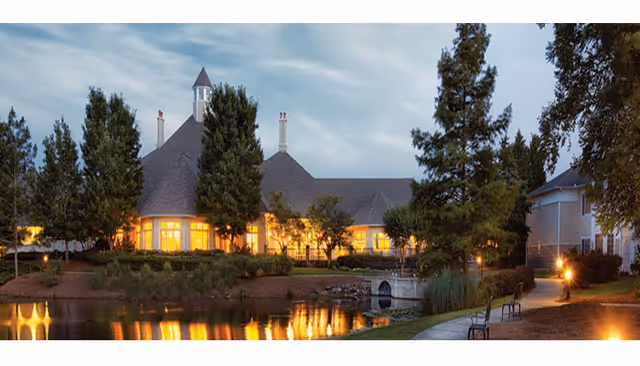 A warmly lit retirement community building at dusk reflected in a pond with a walking path and benches in the foreground.
