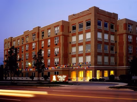 Exterior view of a multi-story brick building at dusk with illuminated windows and a sign in front that reads Senior Suites Of Hegewisch. The building has decorative brickwork and several trees along the sidewalk. Colorful pennant flags are strung along the front of the building.