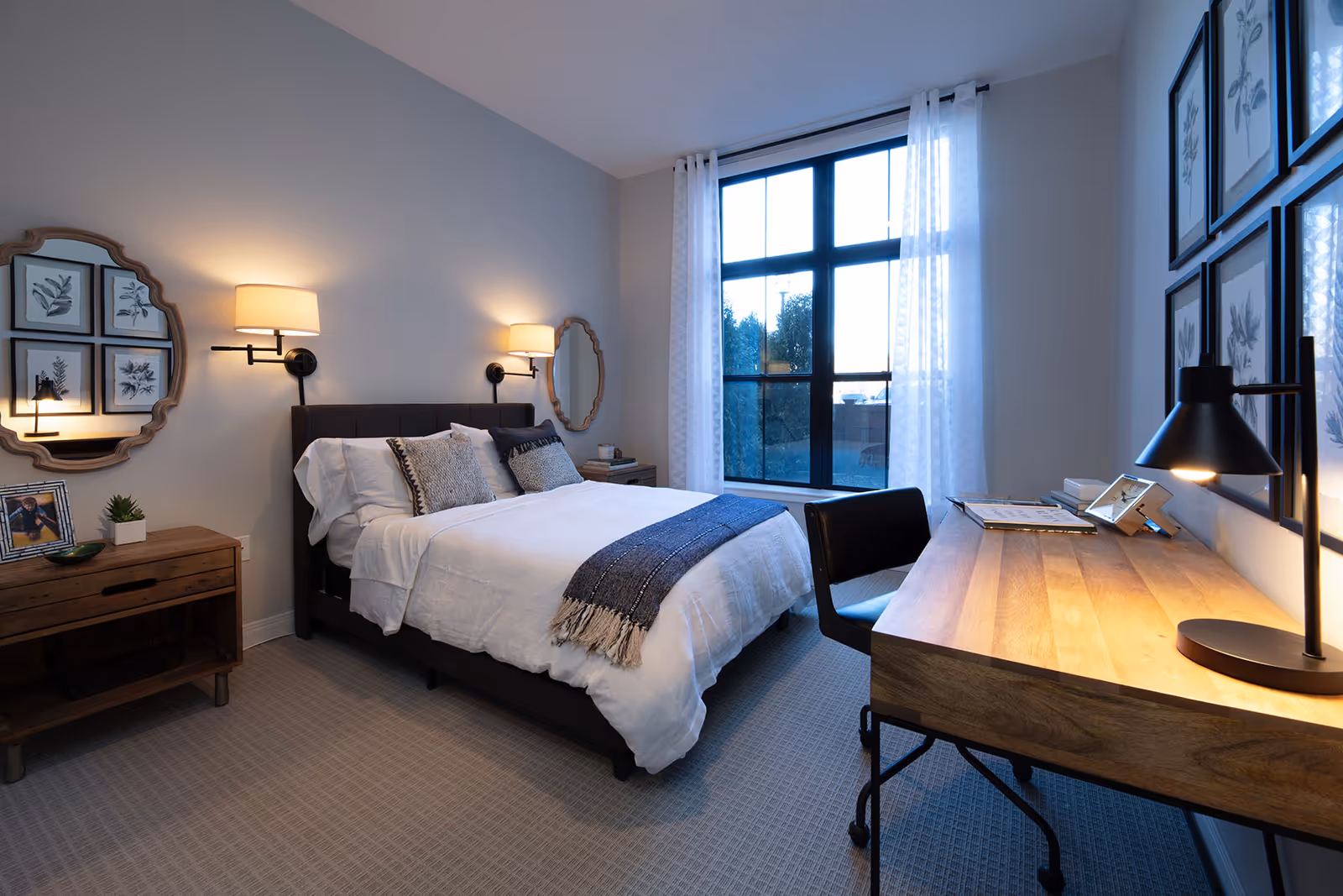 Bright modern bedroom with a made bed, bedside tables and wall sconces, a large window with sheer curtains, and a wooden desk with a lamp.