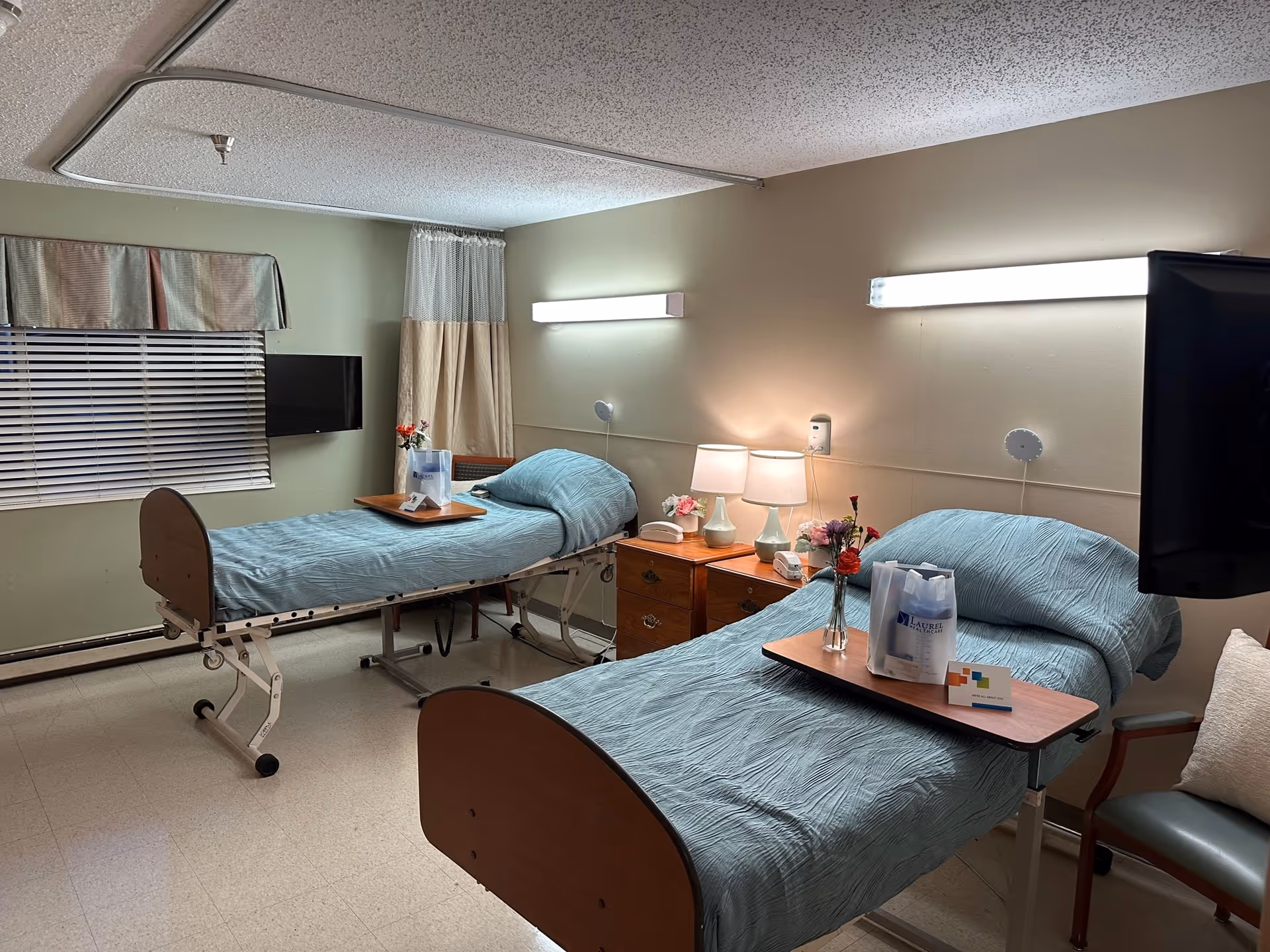 A room with two hospital-style beds, each with light blue bedding and a wooden overbed table. The tables hold flowers and a gift bag. Between the beds is a wooden nightstand with two lamps and a telephone. The room has a window with blinds and a valance, a wall-mounted TV, and a chair with a cushion. The walls are light green and beige, and the floor is tiled.