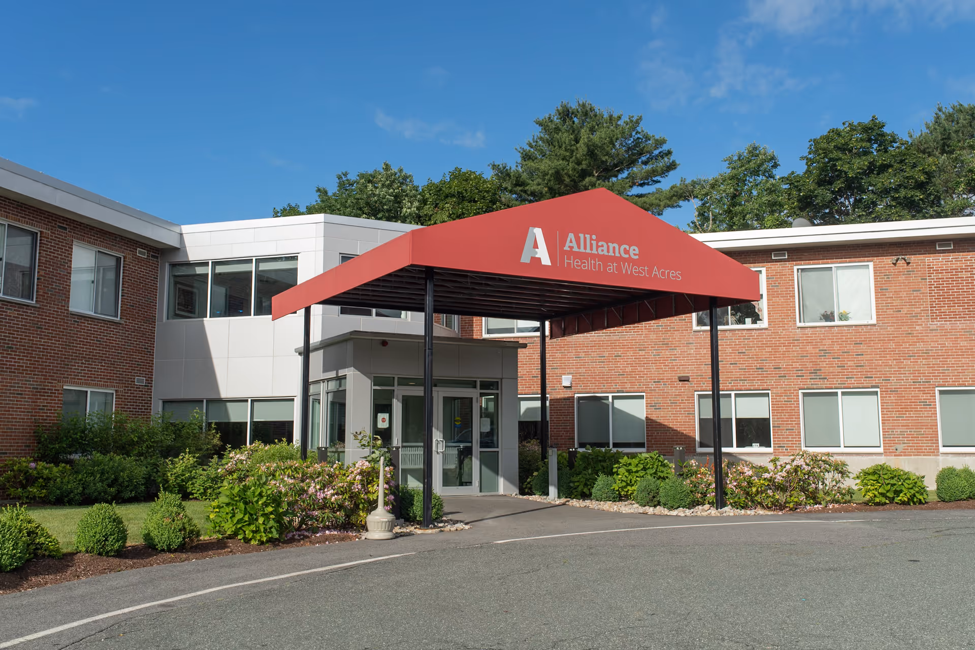 Exterior front view of Alliance Health at West Acres facility showing a brick building with a red canopy entrance and surrounding greenery under a clear blue sky.