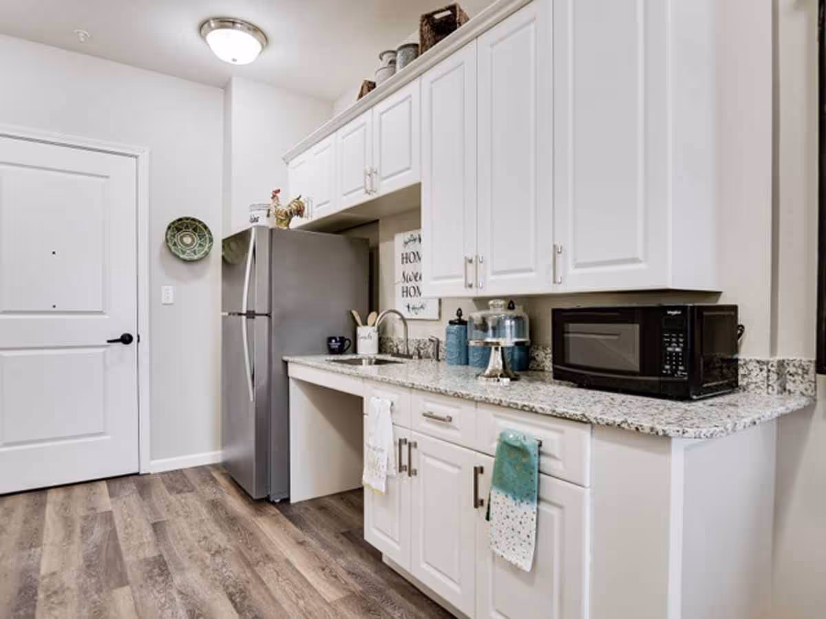 Small kitchenette with white cabinets, granite countertops, a stainless refrigerator, microwave and sink.