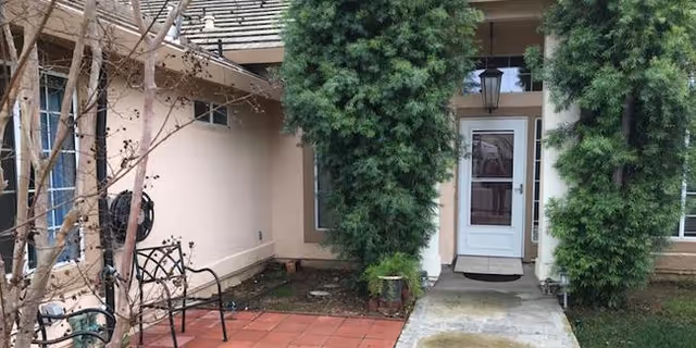 Outdoor view of a residential facility entrance with a concrete walkway leading to a white door flanked by two tall green bushes. There is a small patio area with a metal bench and a leafless tree on the left side.