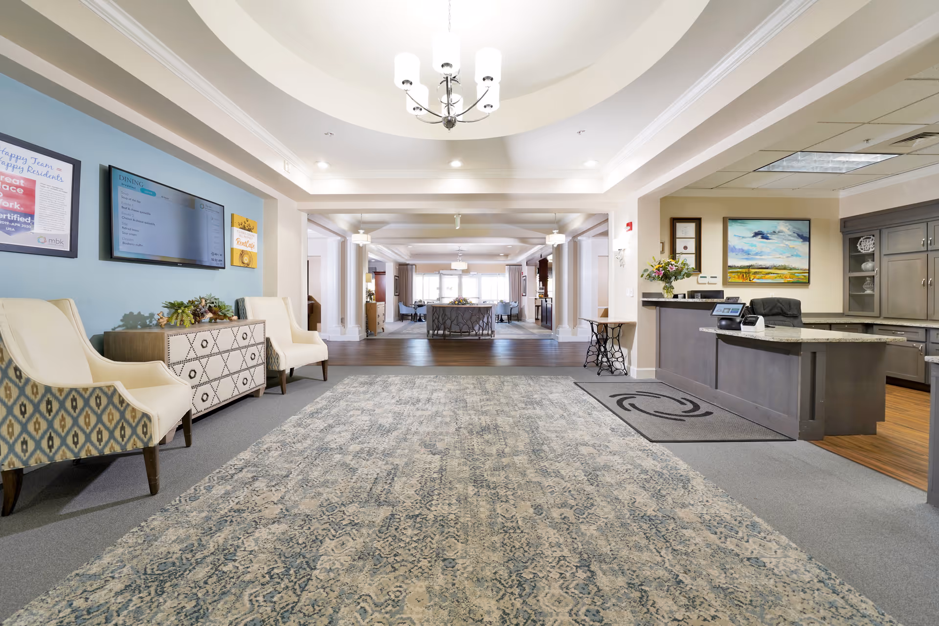 Spacious and well-lit interior area of The Inn at Greenwood Village featuring a large patterned rug, two upholstered chairs, a decorative sideboard, a reception desk with a computer, and a chandelier hanging from a tray ceiling. The background shows an open dining area with tables and chairs.