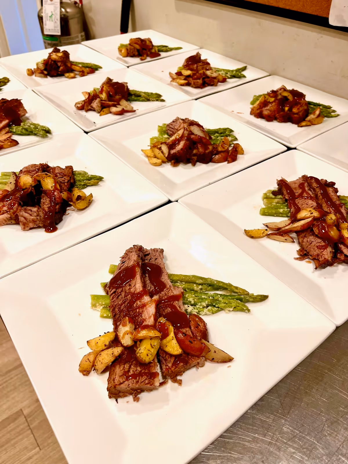 Multiple white square plates arranged on a counter, each with a serving of sliced steak topped with sauce, roasted garlic cloves, and asparagus spears.